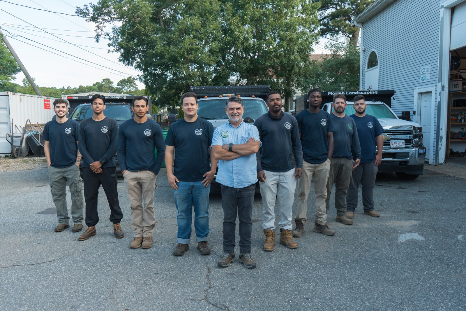 The Moriah Masonry and Landscaping crew of eight with company trucks at the yard in South Yarmouth, MA